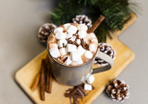 A mug of hot cocoa topped with marshmallows, chocolate pieces and a cinnamon stick rests on a wooden platter decorated with garland, cinnamon sticks and pinecones.